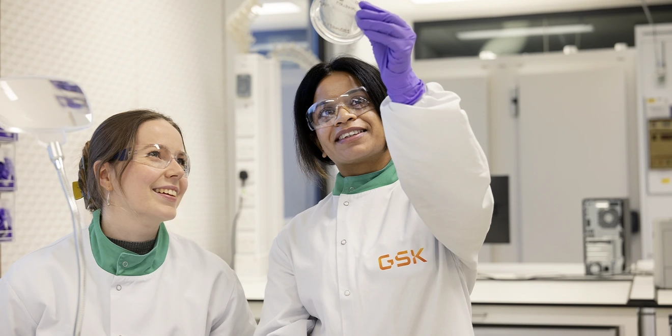Duas cientistas de jaleco branco em laboratório da GSK observando uma amostra.